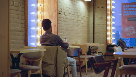student in formal shirt sits alone inside cozy cafe, holding takeaway coffee cup near glowing window lights, with jacket on chair, water bottle on table, and backpacks on bench in softly lit modern space
