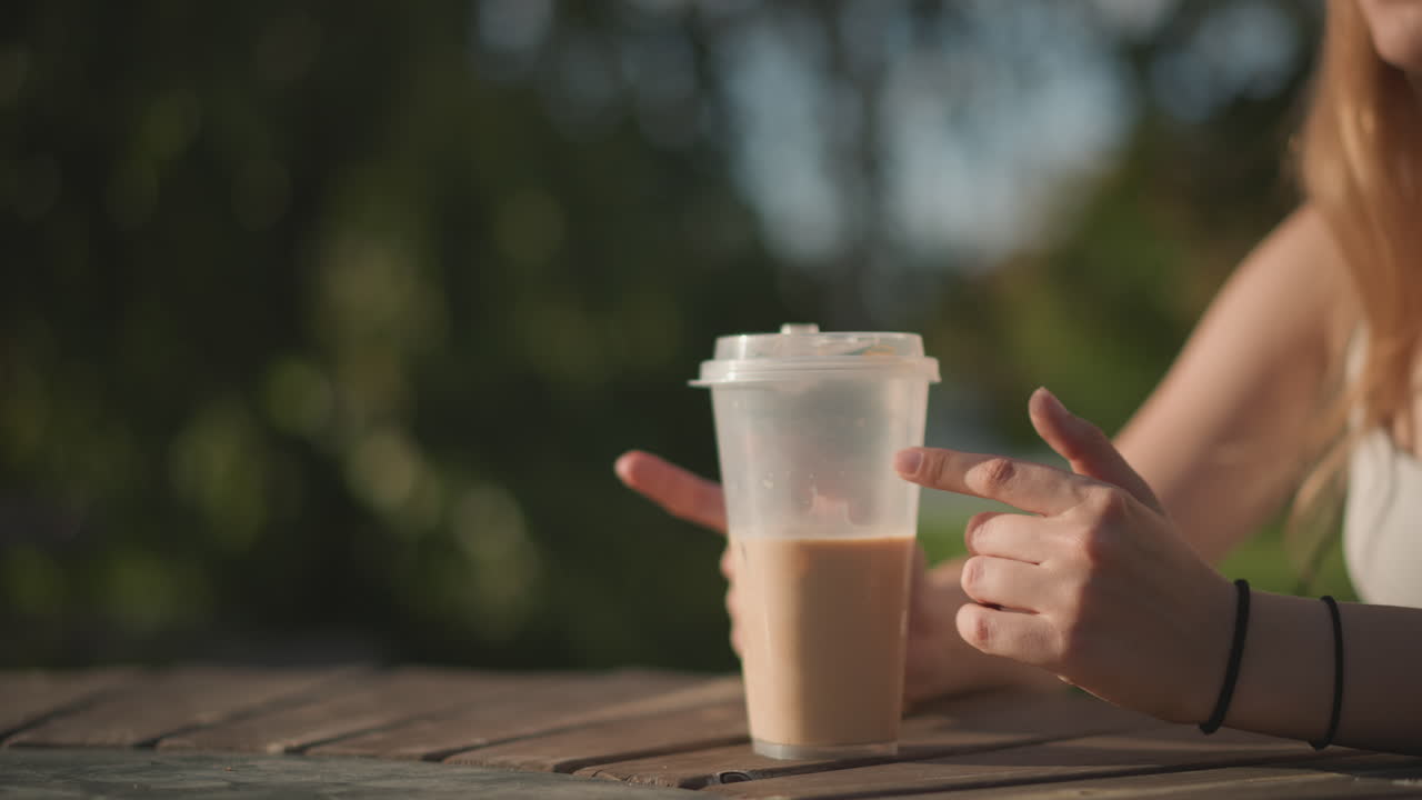 Giovane donna bianca picchietta con la tazza di caffè freddo zuccherato su un tavolo di legno, le dita che scandiscono il ritmo come un ballerino di strada, la luce del sole che filtra tra gli alberi del parco, sfondo bokeh leggero, tonalità estive calde, braccialetto casual