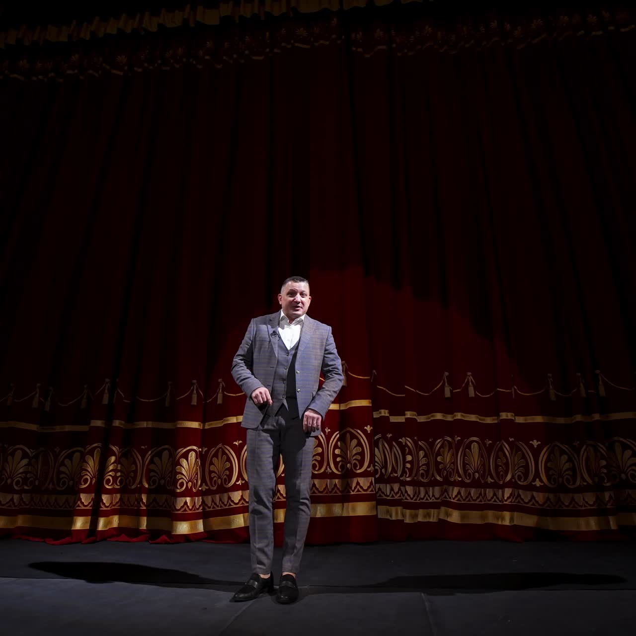 Male host talking on stage. Elegant man in grey costume performing on large scene uses gestures. Actor moving joyfully on stage