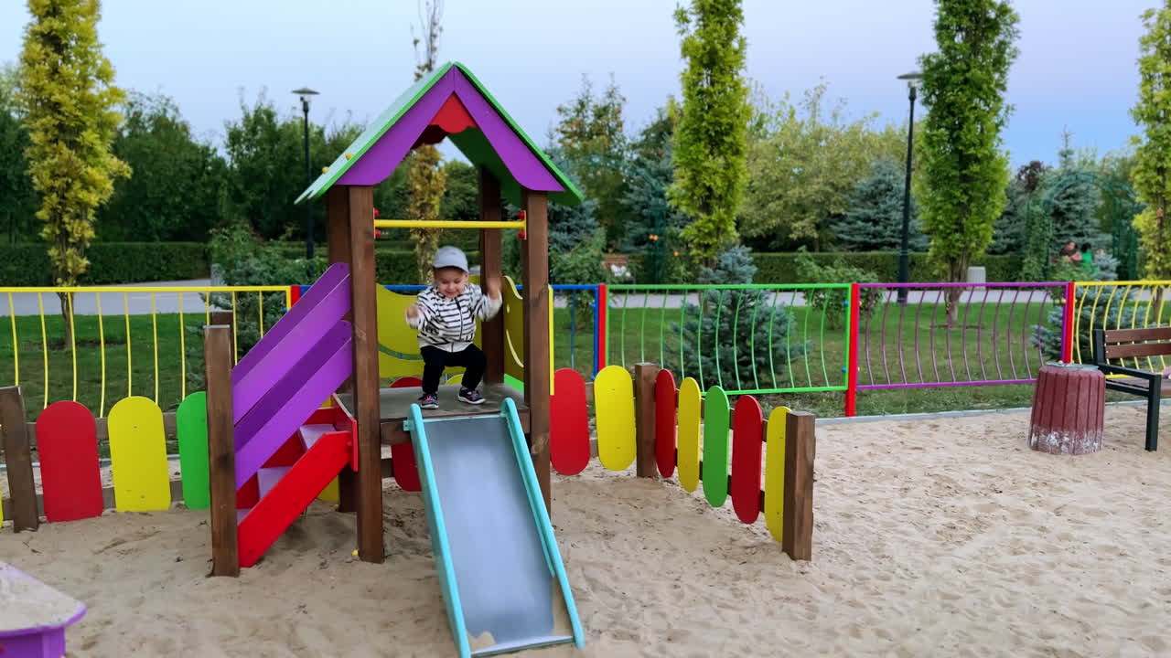 Smiling baby boy in stripy bomber jacket is on the slide. Kid tapping feet by the slide and then goes down.