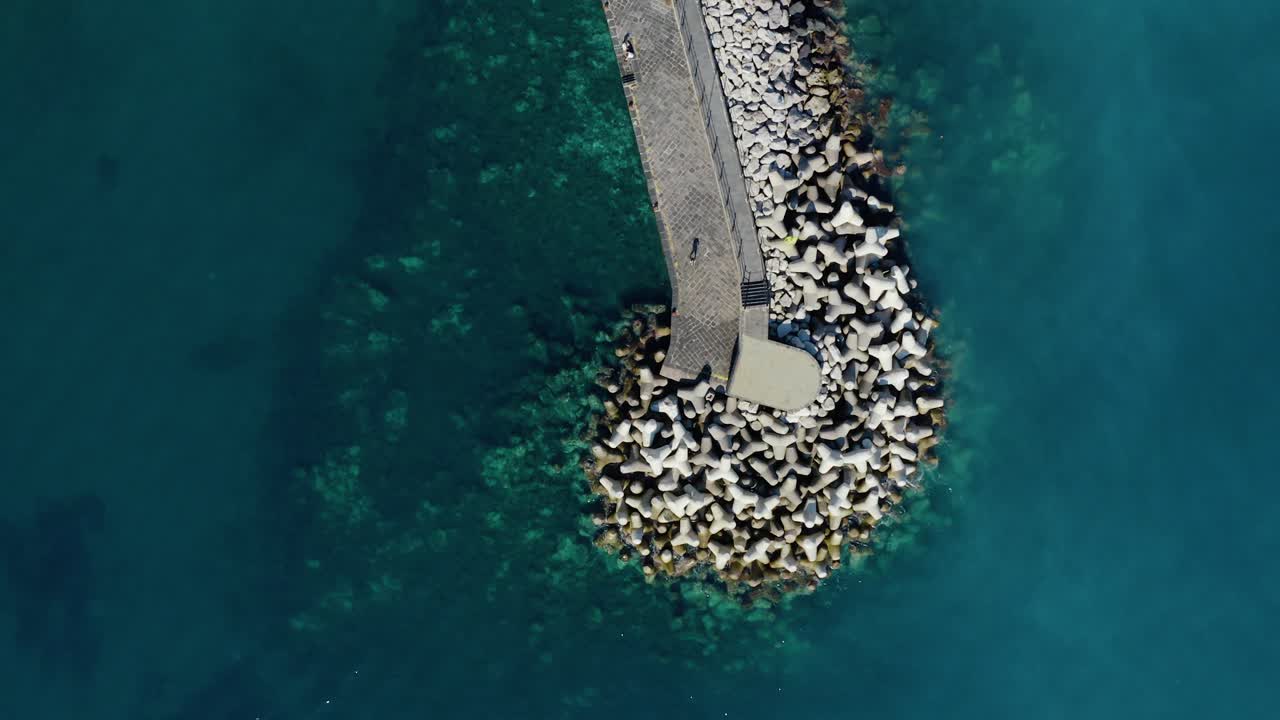 Vista aerea dall'alto con drone di un molo frangiflutti in pietra curvo che si estende in un mare turchese e cristallino con barriere frangiflutti costiere
