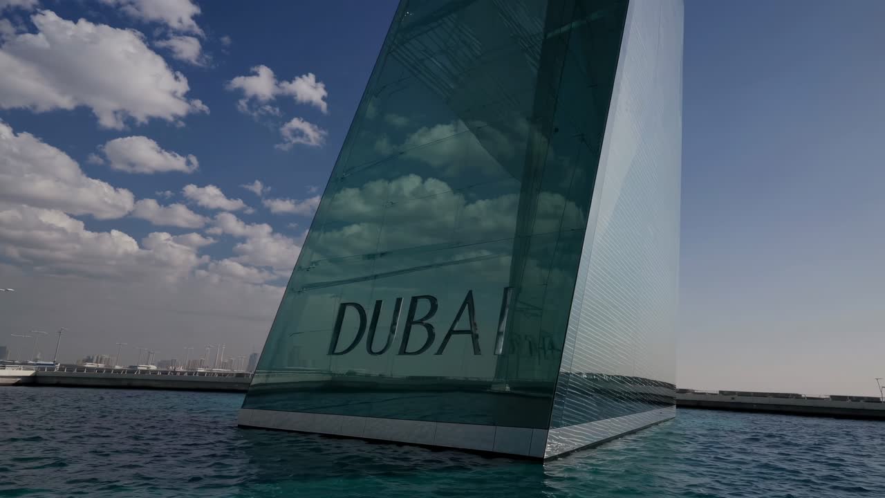 Glass wall featuring the Dubai logo reflects fluffy clouds under a bright sun, with a stunning blue sky and serene sea, capturing the essence of modern architecture in Dubai, UAE