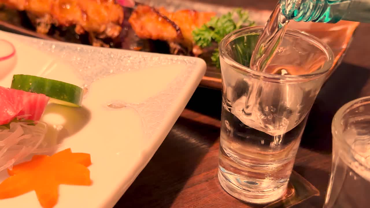 Sake limpido si versa in un bicchiere accanto a sashimi e maki sotto una calda illuminazione da ristorante.