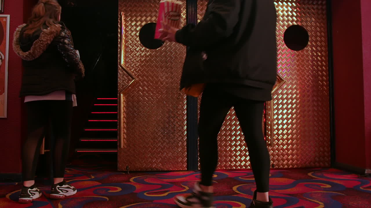 A mother and daughter entering a cinema theater holding a tub of popcorn, through metallic double doors entrance with circular windows.