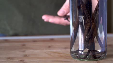 close up person drops vanilla pods into glass bottle of clear alcohol