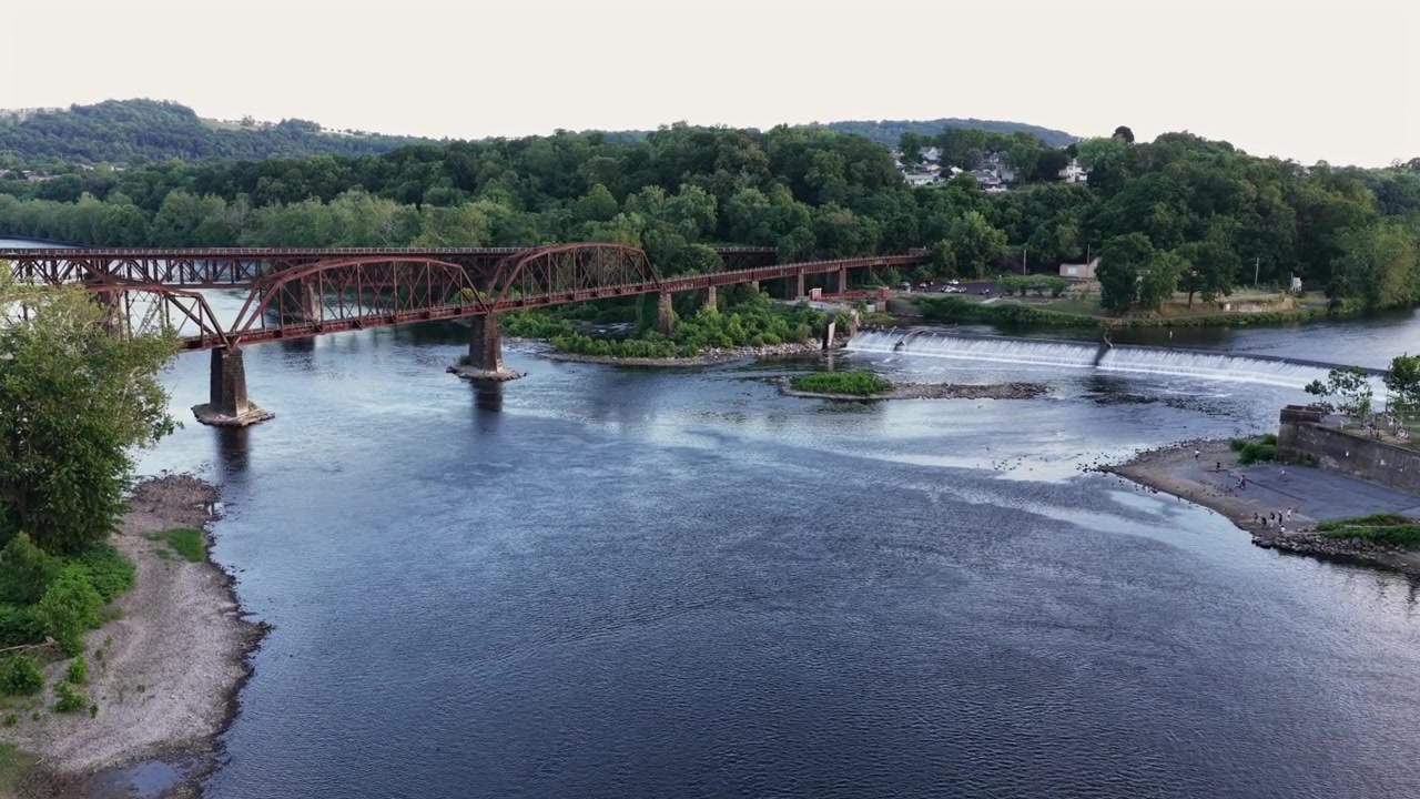Ponte ferroviario a traliccio in acciaio rosso arrugginito che attraversa un ampio fiume vicino a Easton, Pennsylvania; verdi colline boscose si ergono oltre una piccola isola alberata e rapide rocciose a valle. Ampia ripresa aerea con drone.