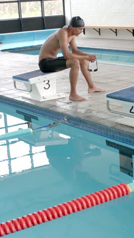 vertical video: swimmer sitting on poolside bench holding goggles, preparing for swim training