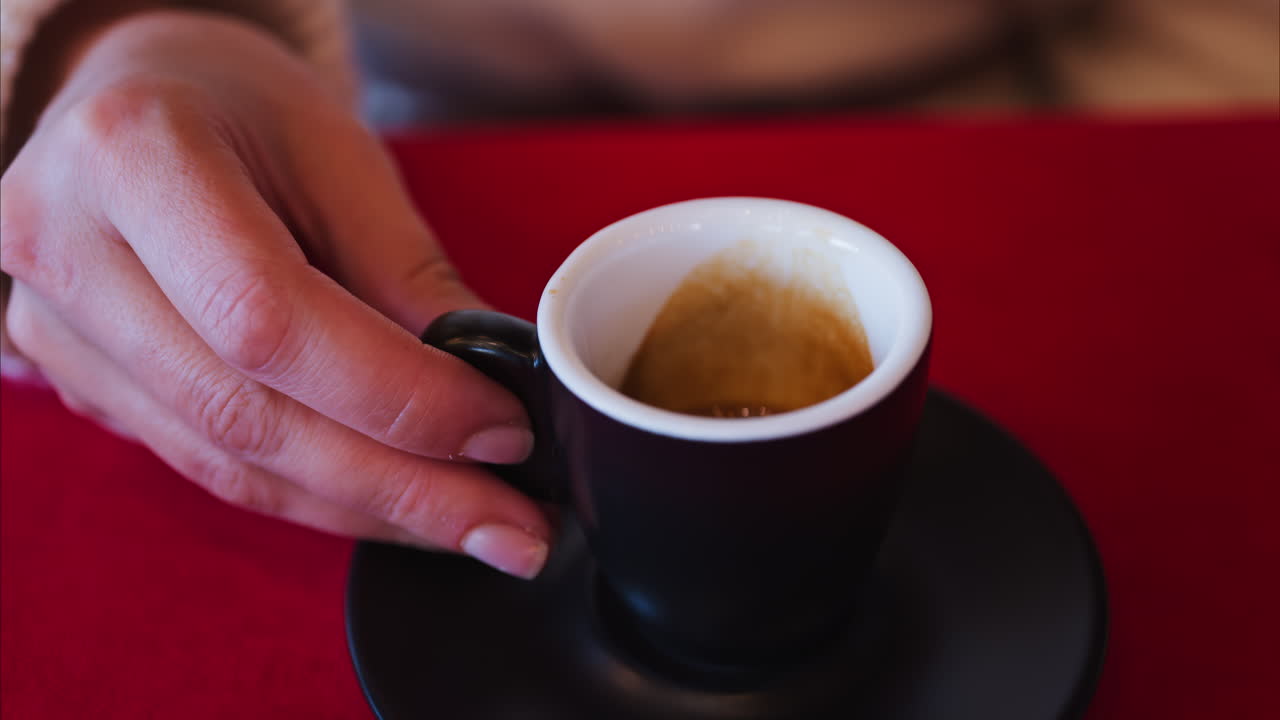Close up of a woman holding an espresso in a black cup on a red tablecloth at a restaurant