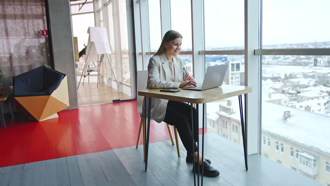 Young woman at her workplace in light spacious office. Business lady sitting at desk and working at laptop. Cityscape at the backdrop.