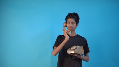 Handsome-shaven-Indian-young-guy-20s-in-black-t-shirt-isolated-over-blue-background-in-studio-with-old-vintage-telephone