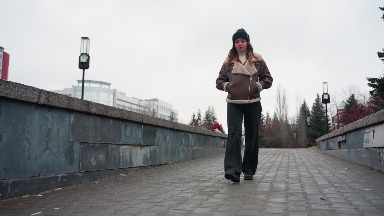 Thoughtful girl in black cap brown jacket and trousers walking slowly hands in pockets on paved pathway surrounded by pine trees and urban buildings under cloudy sky during cold peaceful autumn day