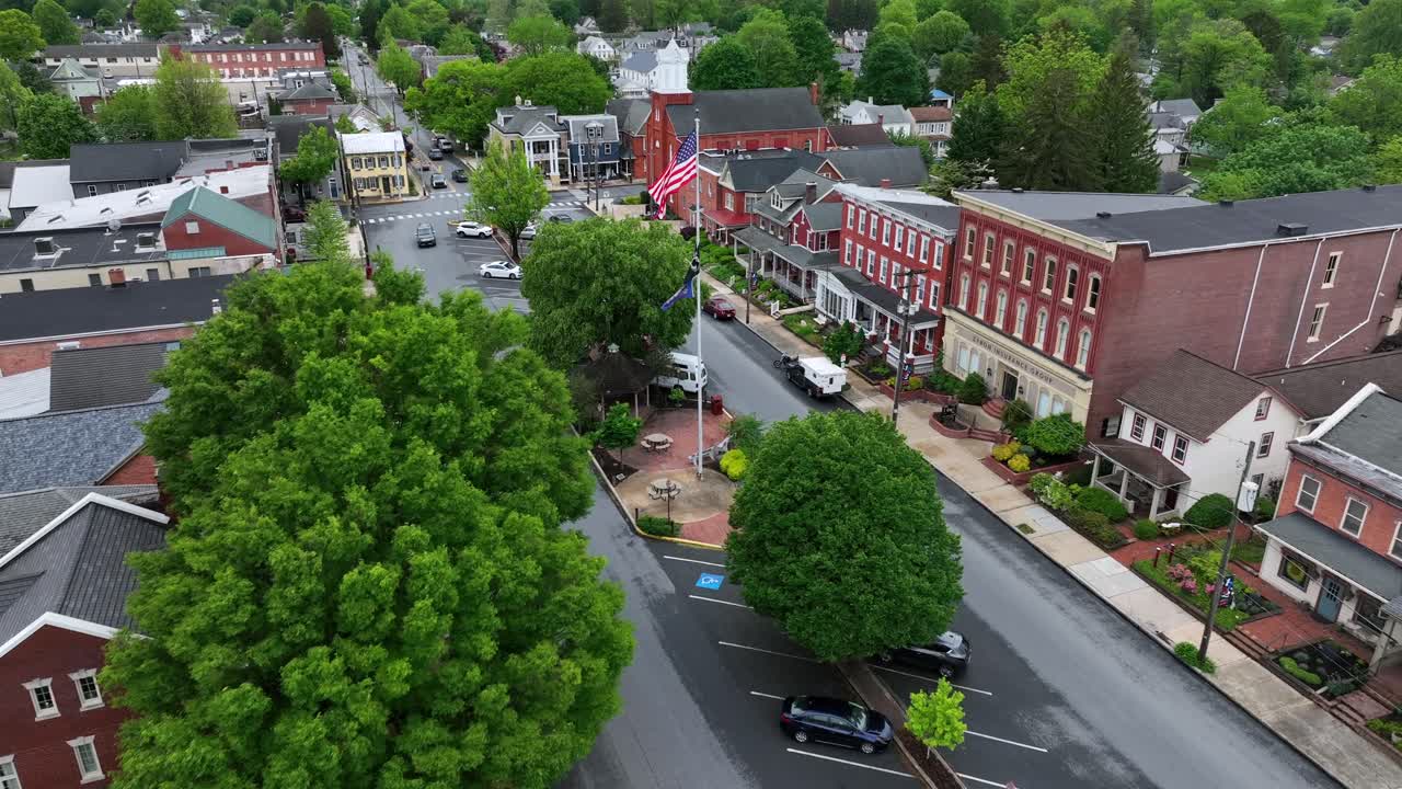Patriotic American flag waves over small town in USA. Main Street in Small Town America square at cloudy day in spring. Aerial Approaching shot. Manheim, Pennsylvanias.