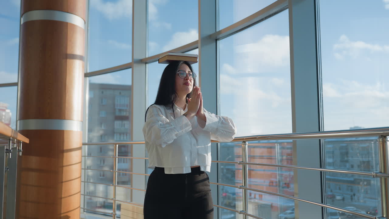 Woman balancing book on head while walking inside sunlit glass corridor, lifting hands gently as she walks with confidence and joy, snowy urban landscape visible outside through large windows