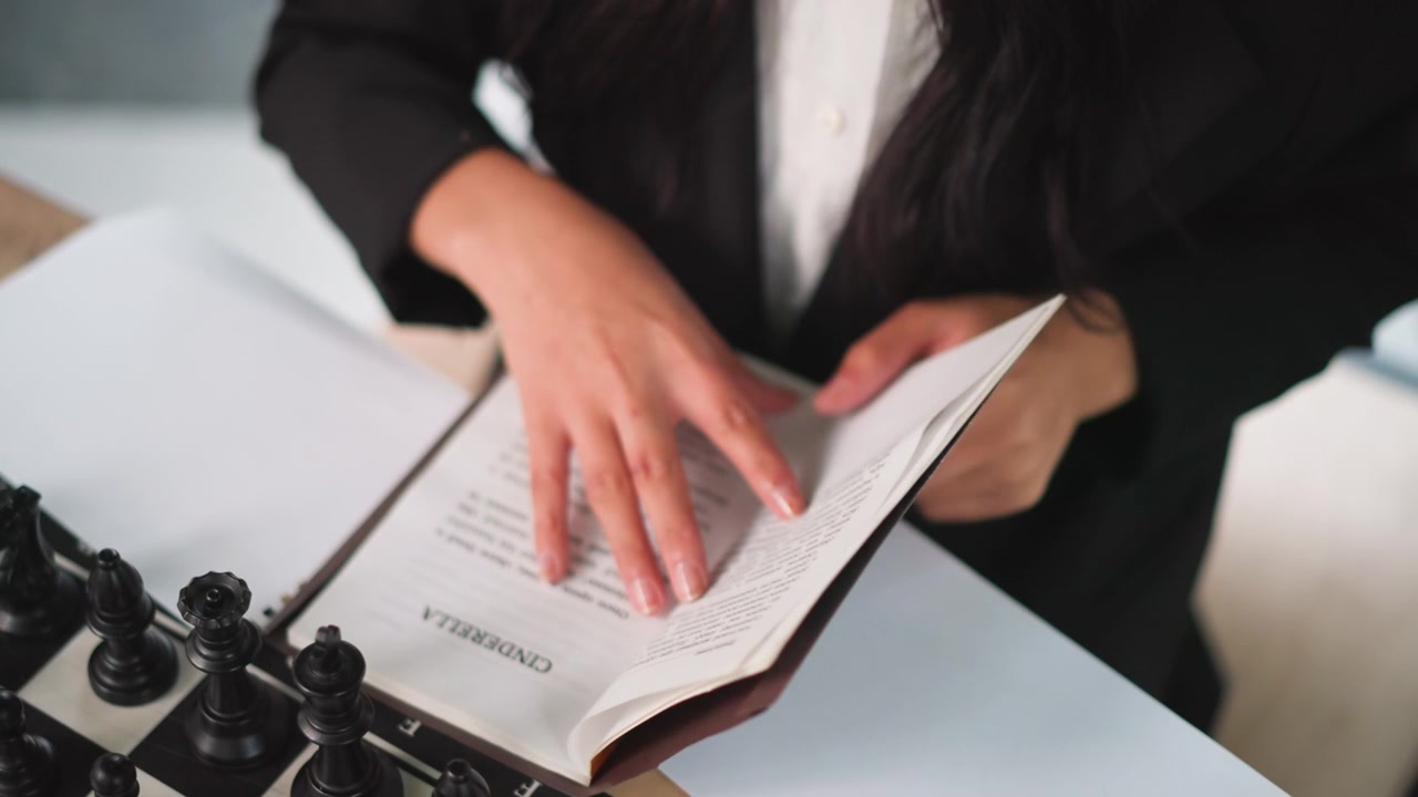 Primo piano di una donna in abbigliamento professionale che legge un libro aperto intitolato Cenerentola con pezzi degli scacchi neri sul tavolo, concentrandosi sulle sue mani e sul testo. La scena è al chiuso sotto una luce soffusa che enfatizza