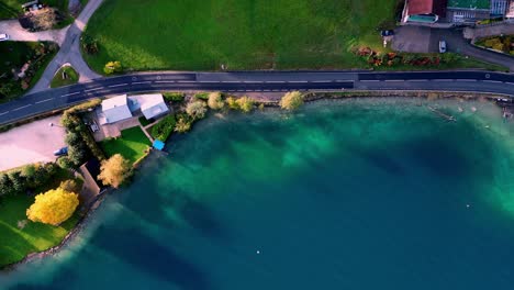 Soar-above-a-winding-road-through-emerald-trees,-reaching-a-shimmering-blue-lake