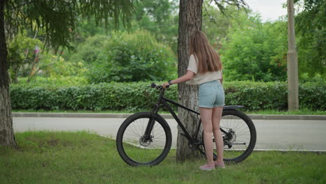 woman in jean shorts approaches bicycle leaning against tree in serene park, removes stand, and begins moving with bike, background of lush greenery and paved path