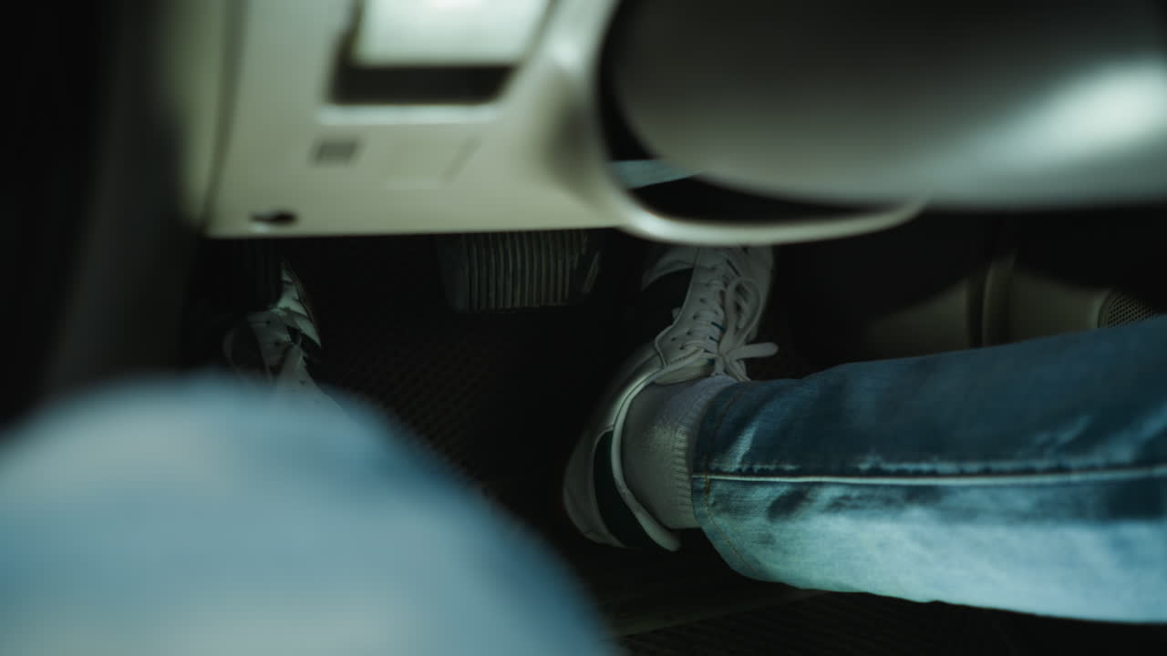 Primer plano de un pie en el pedal dentro de un coche; persona que viaja al trabajo en vaqueros quitando el pie del freno y pisando el acelerador; zapatilla informal sobre la alfombrilla texturizada; luz tenue en el habitáculo; salpicadero difuminado; tensión en el trayecto urbano; estudiante