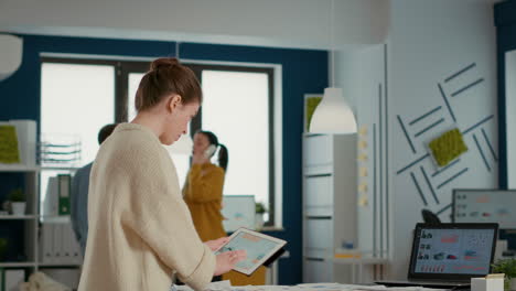 woman holding digital tablet zooming in on sales pie chart and comparing data with statistics on laptop screen and papers