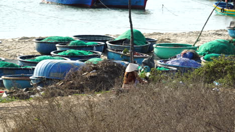 Vietnamese-woman-with-child-riding-motorbike,-beach-with-coracle-boats