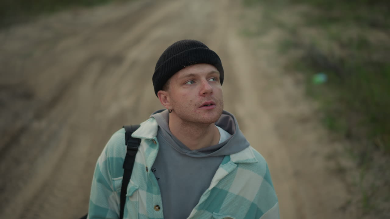 Primer plano contemplativo de un excursionista caucásico fumando en un camino de tierra con gorro y mochila, mirando al cielo mientras inhala un cigarrillo en un ambiente melancólico al atardecer, con un sendero forestal de fondo y una pausa de aventura tranquila.