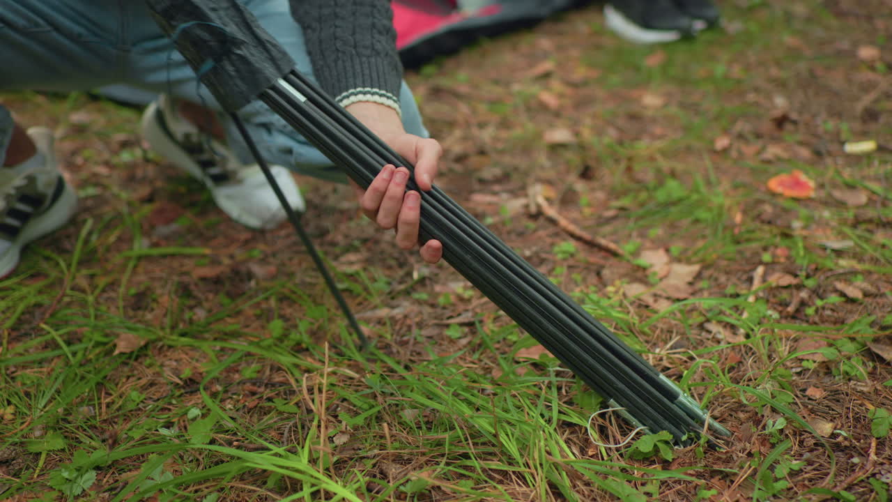 Close up of person squatting on forest ground removing tent poles from bag as one pole slips and is picked up, with leg view of another person standing nearby on grass covered soil