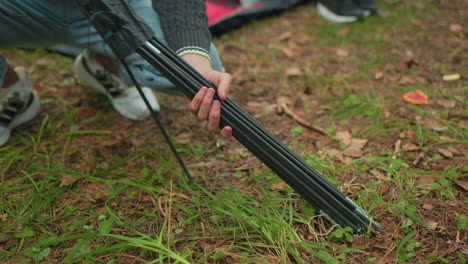 close up of person squatting on forest ground removing tent poles from bag as one pole slips and is picked up, with leg view of another person standing nearby on grass covered soil