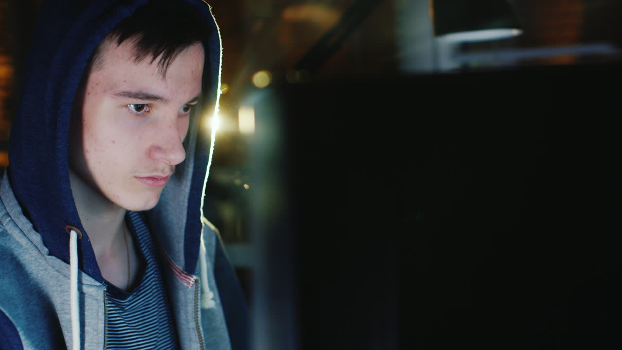 후드를 쓴 십대가 어둠 속에서 컴퓨터 모니터 앞에 앉아 있다 1