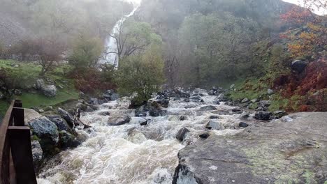 Slow-motion-powerful-rapid-flowing-water-cascades-under-wooden-bridge-from-woodland-valley-waterfall