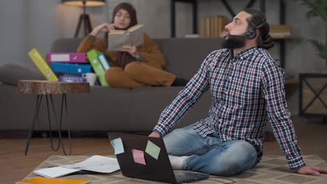 wide shot portrait of busy serious overworking middle eastern man in earphones typing on keyboard analyzing graph sighing and closing laptop. overworked handsome guy in home office wife at background.