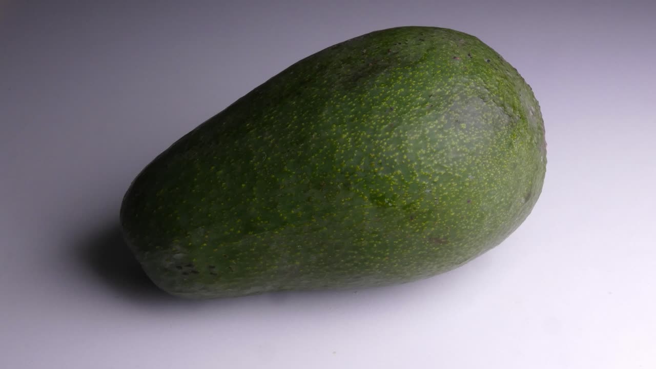 Un avocado verde peruviano maturo su uno sfondo bianco con una fonte di luce che si muove sopra di esso, facendo muovere le ombre.
