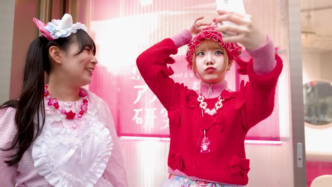兩位身穿鮮豔粉紅色Decora街頭風格的女士，正在東京繁忙的街道上自拍。