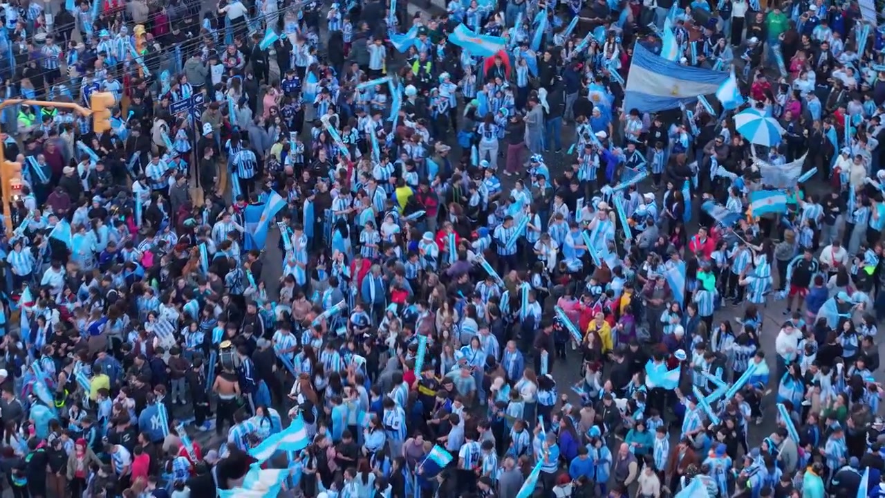 Imágenes aéreas de dron capturan una multitud vibrante ondeando banderas argentinas, celebrando una victoria de fútbol en la Plaza 25 de Mayo, San Juan, Argentina. De día, ambiente festivo, vista cenital.