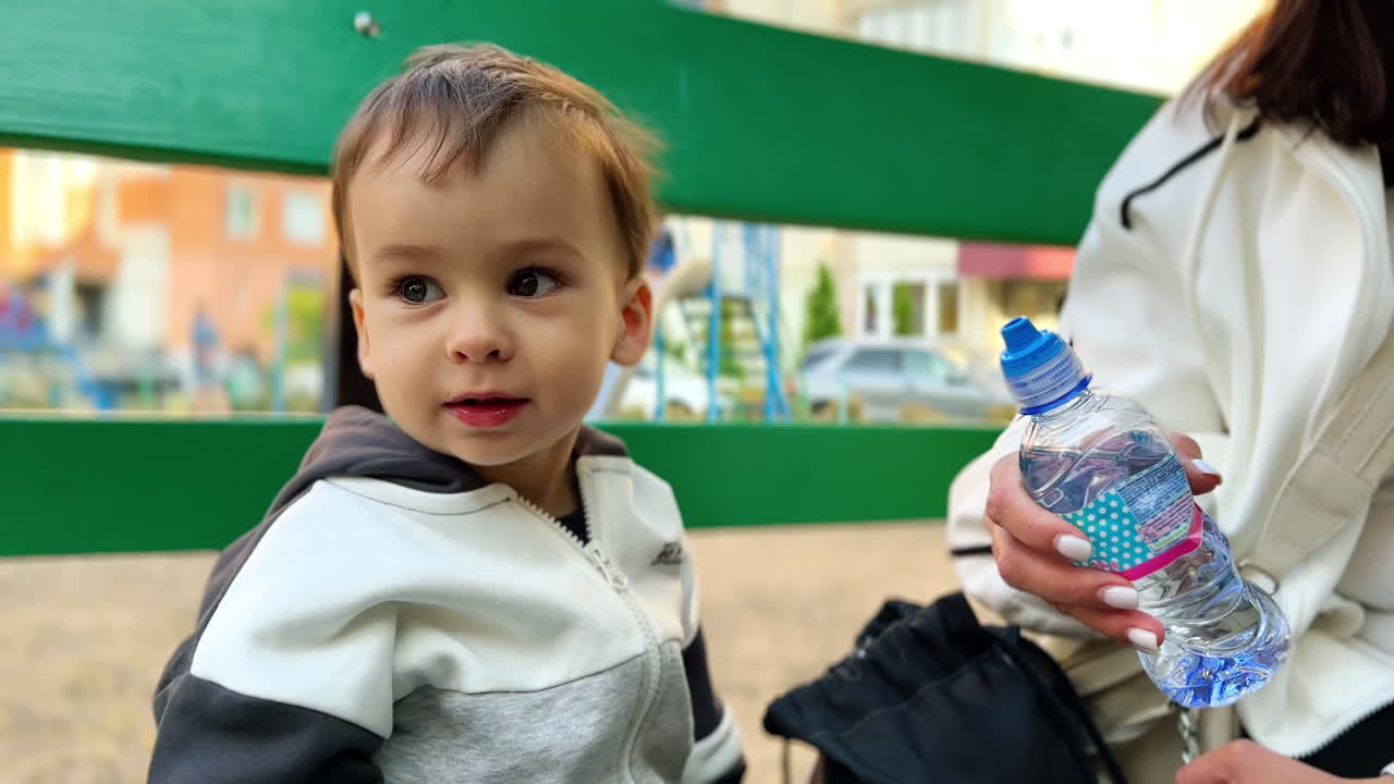 Mom's hand holding water bottle for her baby boy. Kid drinks hungrily and then points at something aside with a finger. Close up.