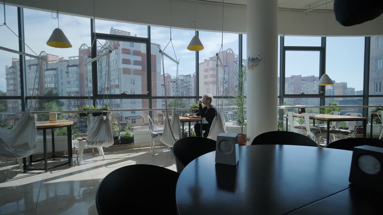 Giovane uomo con i capelli biondi che gusta un panino e un caffè freddo a un tavolo di un caffè vicino a una finestra con vista sulla città, luce naturale e piante in vaso. Scena di ristorazione rilassata e informale