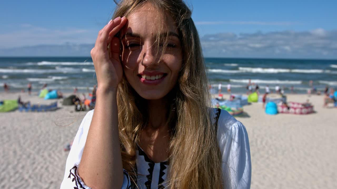 A close-up of a woman smiling at the beach with her hair blowing in her face and other people sitting on the sand in the background