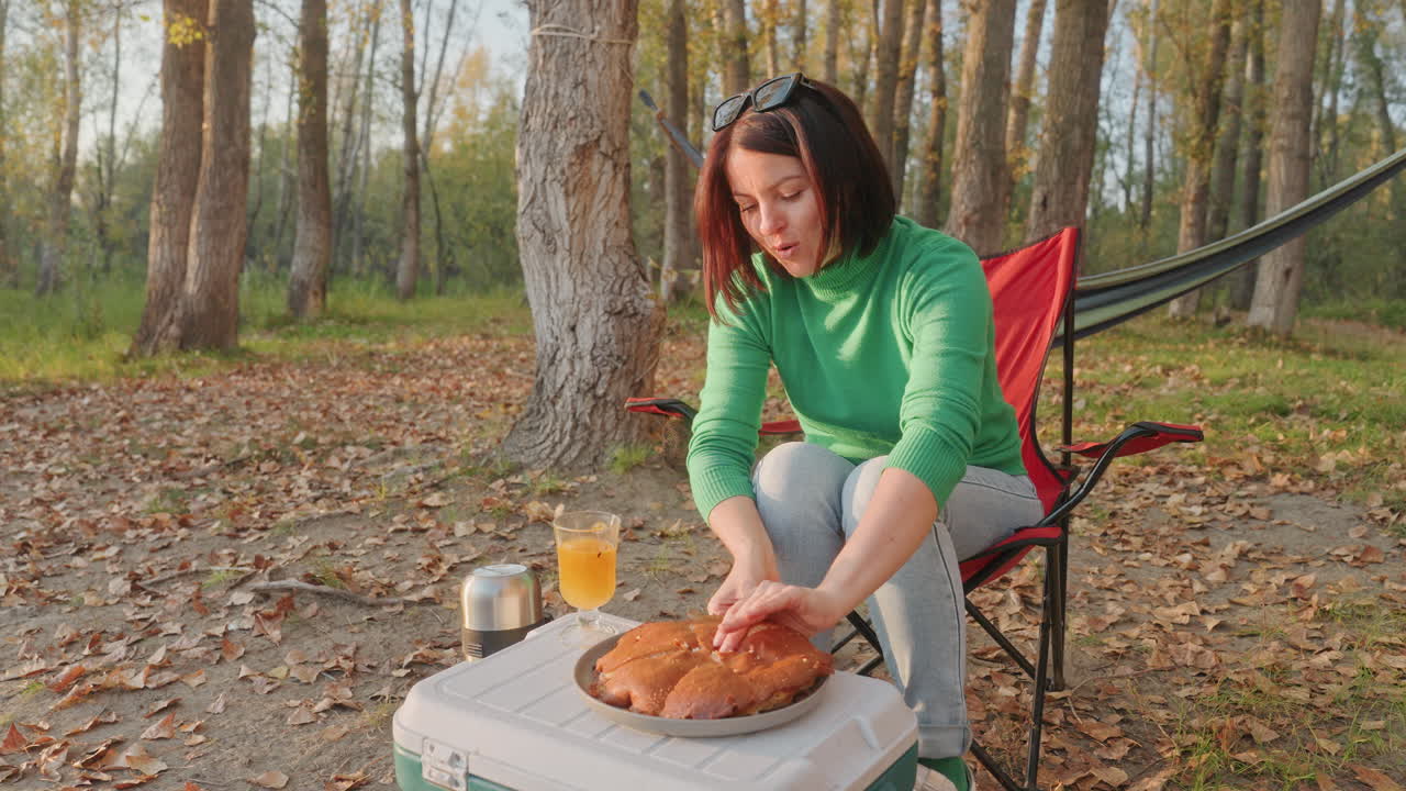 Kvinde i skovens campingstol river brød. Hyggelig campingmorgen med hjemmebagt brød på kølebord, kedel og glas juice ved siden af, hængekøje spændt ud mellem træer, nedfaldne blade under fødderne, blødt