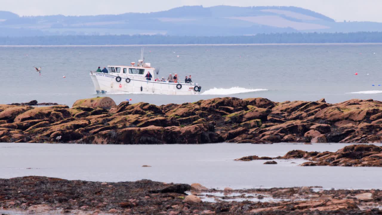 A white tour boat travels along a rocky coastline under daylight, with distant hills and calm sea, captured in a steady lateral camera pan