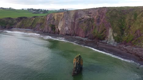 drone shot of sea stack in emerald green sea with waves lapping on a beach with red cliffs at copper coast waterford ireland