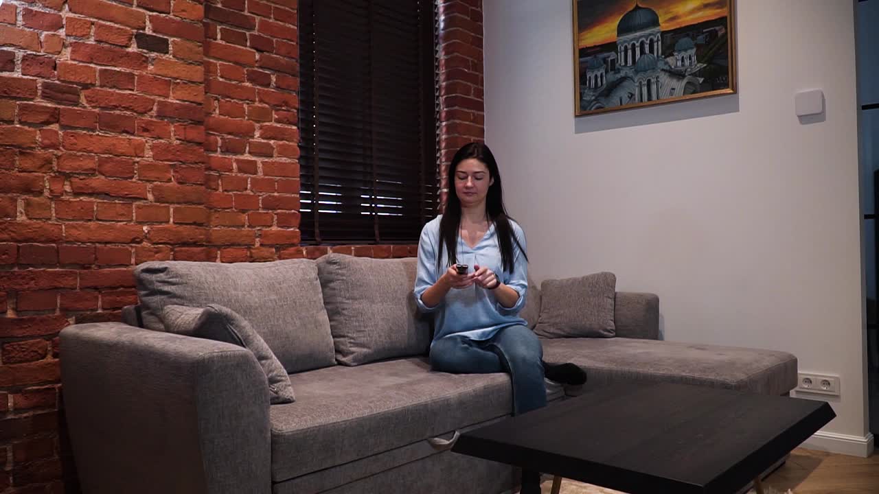 una mujer se sienta en el sofá, enciende la televisión con un control remoto y mira televisión, y se relaja en la sala de estar de su casa con una pared de ladrillo rojo