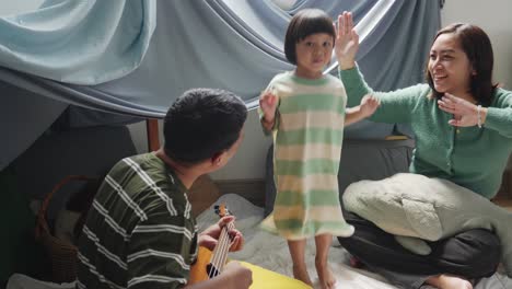 happy asian family, young father playing guitar while mother and daughter singing and dancing together at home