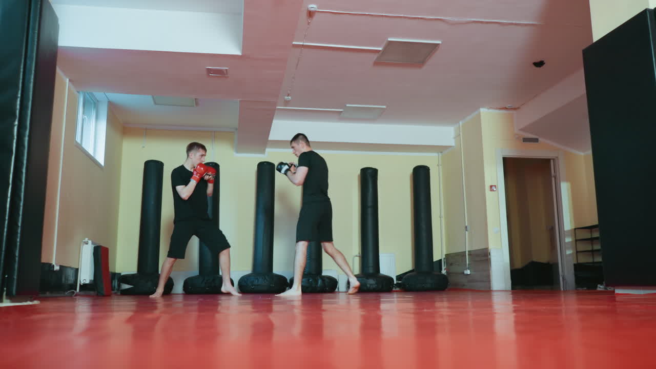 격투기 체육관에서 스파링 연습을 하는 운동선수들의 모습이에요. 한 명은 펀치를 날리고 다른 한 명은 방어 자세를 취하고 있어요. 두 명 모두 검은색 운동복과 장갑을 착용하고 빨간색 매트 위에서 펀칭백에 둘러싸여 훈련하고 있어요.