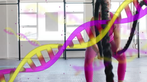 animation of dna strands over happy caucasian woman cross training with battle ropes at gym