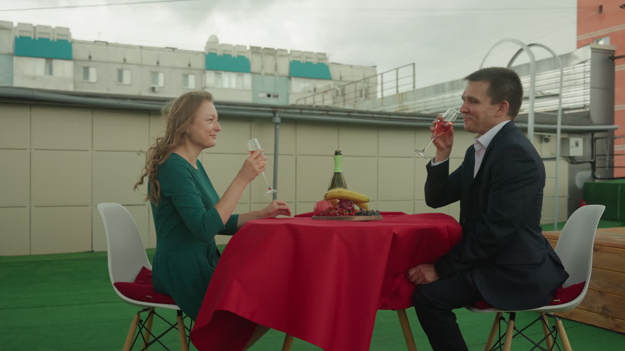 Legende date på en tagterrasse med byens brise. Hvidt ingeniør- og kunstnerpar griner, mens de skåler med vin og druer, vindblæst hår, rød dug, hængende gyngestol, afslappet aftenstemning, lyst og ærligt.