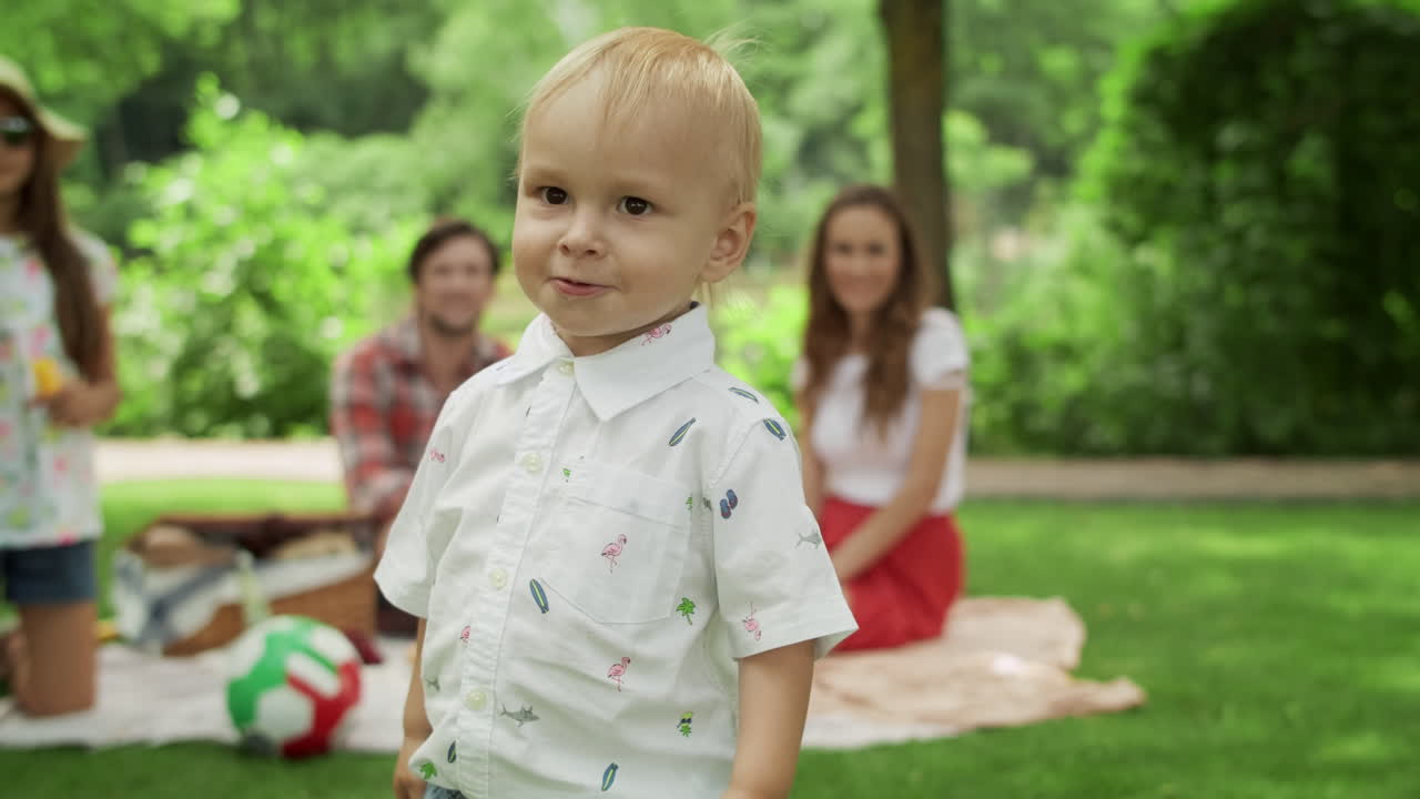 Blonde toddler covering face with hands. Family sitting on blanket in park.
