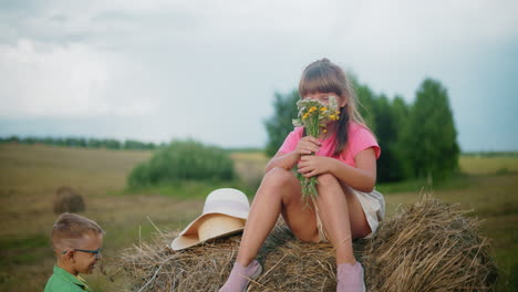 little girl sits on hay bale playfully covering face with flowers while younger brother laughs at her, a sun hat rests beside her as they enjoy a lighthearted moment in open countryside