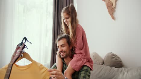 una pequeña chica morena en un vestido rosa se sienta en los hombros de su padre, un hombre moreno que está vaporizando su vestido amarillo en un apartamento moderno. un padre soltero se comunica con su hija mientras vaporiza un vestido amarillento en casa en un departamento moderno