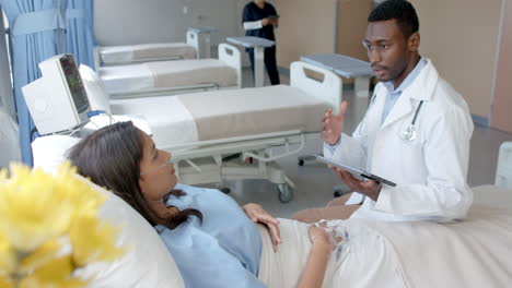 Diverse-female-patient-in-hospital-bed-and-male-doctor-with-tablet-talking-in-ward,-slow-motion