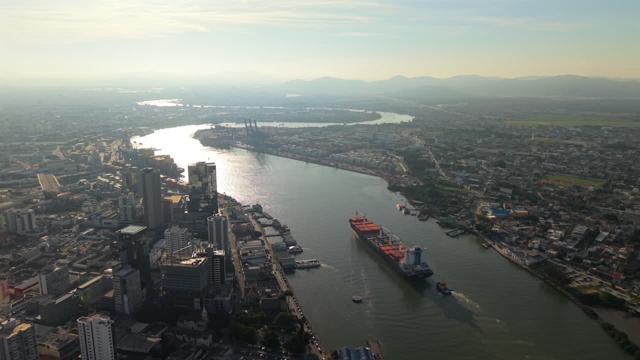 A majestic panoramic aerial shot of a large cargo ship entering the Port of Itajaí, Brazil, during a hazy, beautiful golden hour. A cinematic view of global commerce