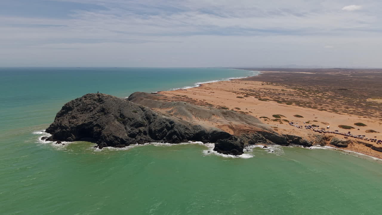 Filmagem espetacular de drone do Pilón de Azúcar em Cabo de la Vela, Colômbia, mostra falésias desérticas áridas encontrando ondas caribenhas turquesa em plena luz do dia, capturando beleza rústica e paisagem dramática