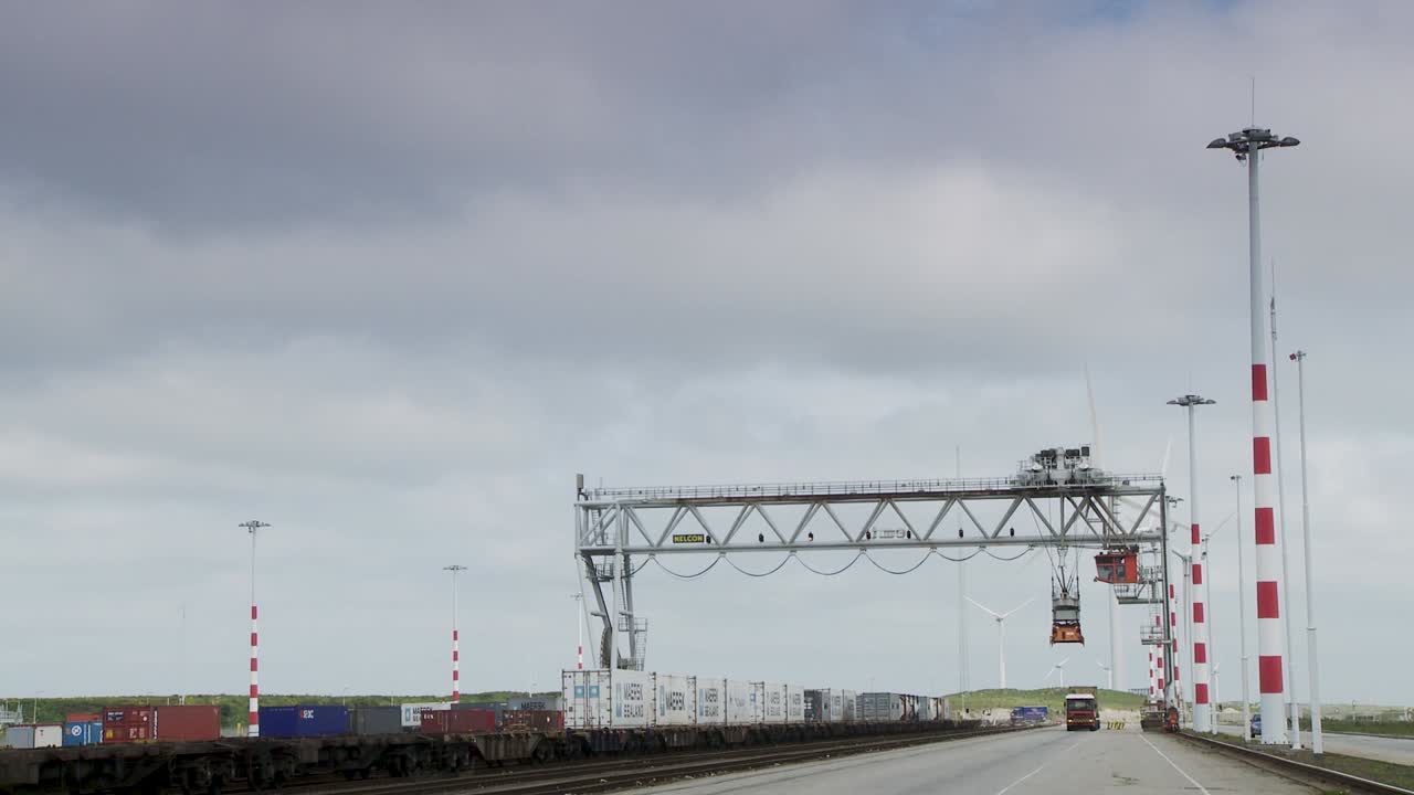 Freight train passing under a gantry crane at a container terminal, overcast sky